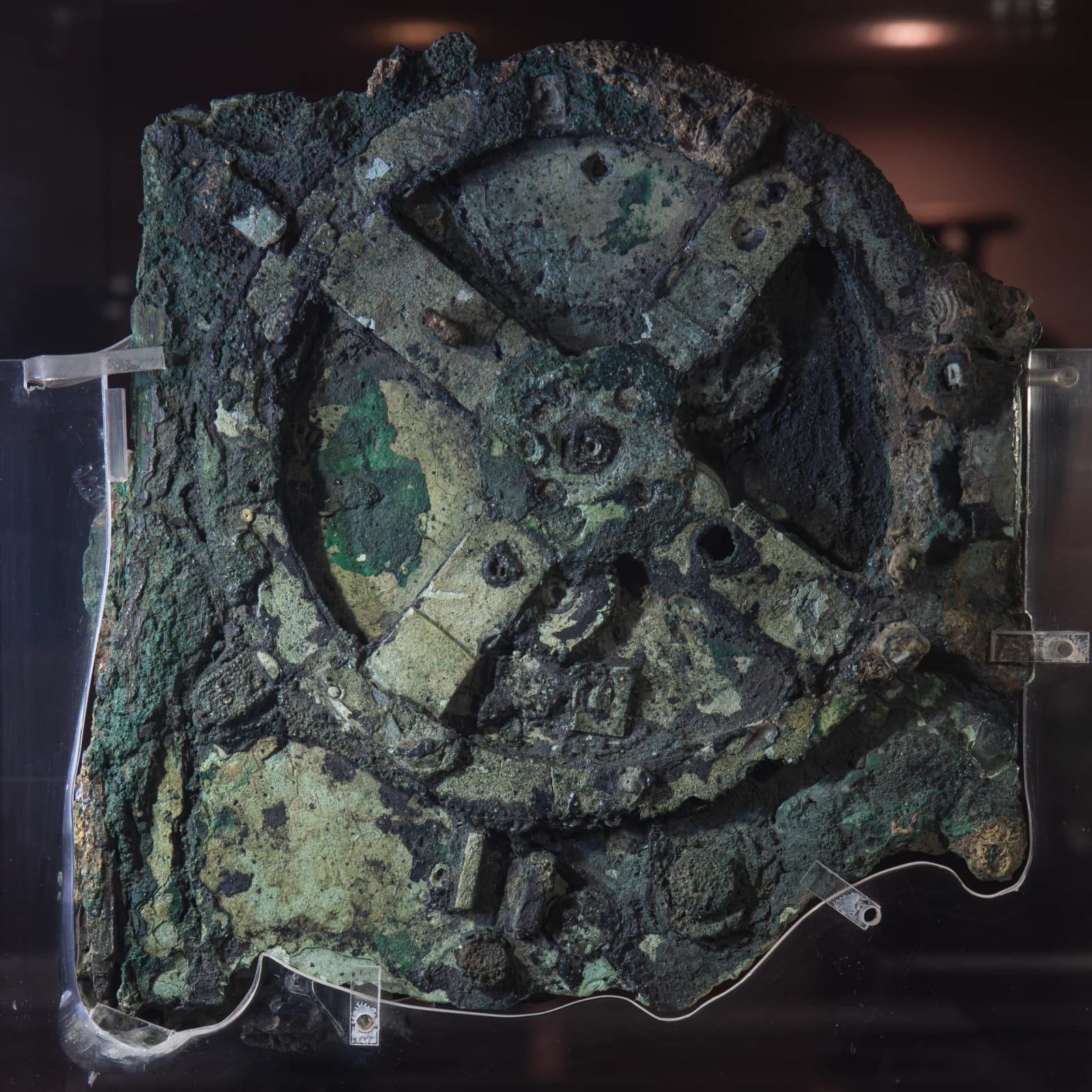 The Antikythera mechanism, fragment A · Wikimedia Commons · CC BY 4.0 A corroded, greenish lump of bronze in which the outline of a large toothed wheel with four spokes can still be seen.