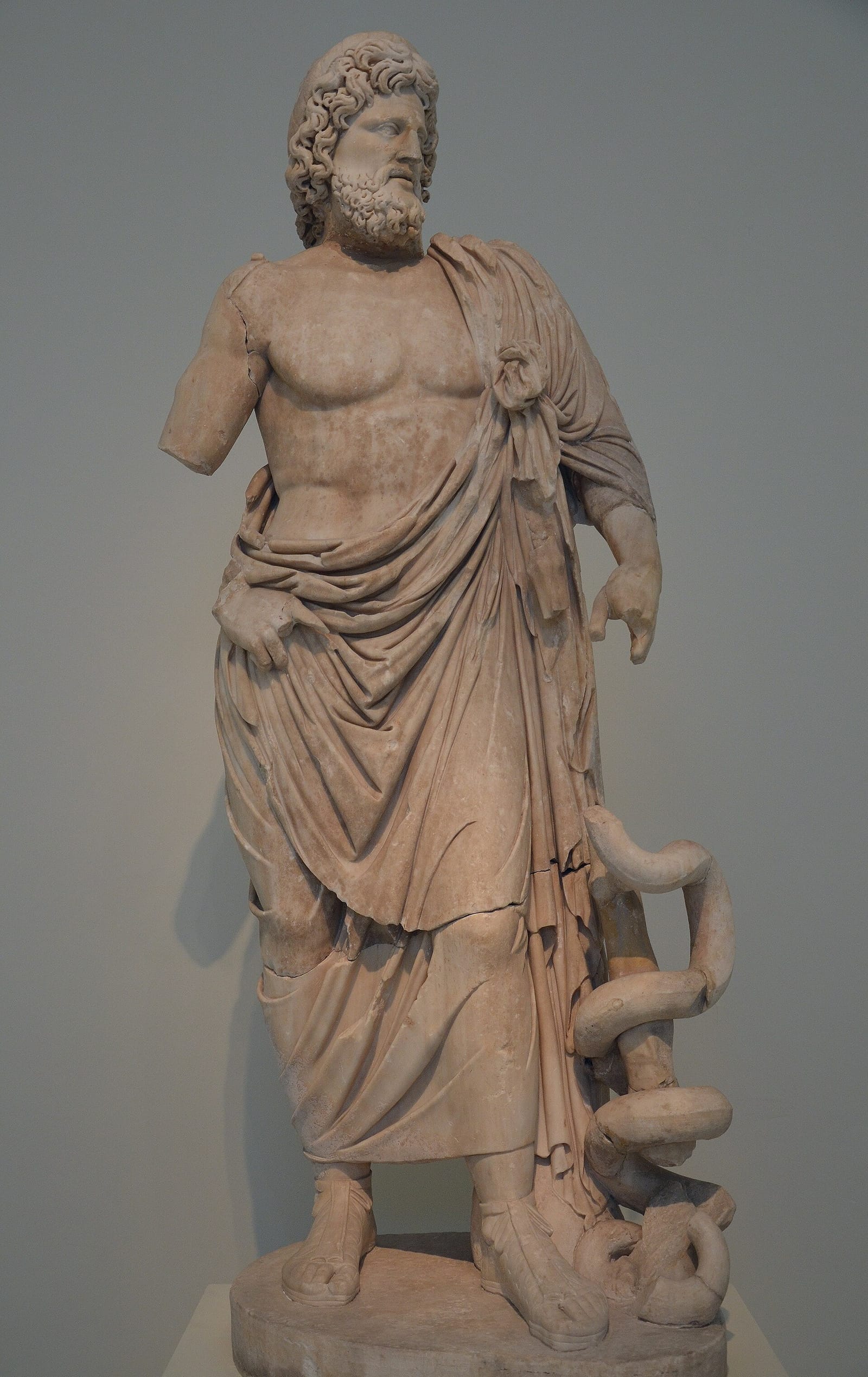 A marble statue of a bearded god in a cloak draped over one shoulder, a snake coiled around the staff at his side.