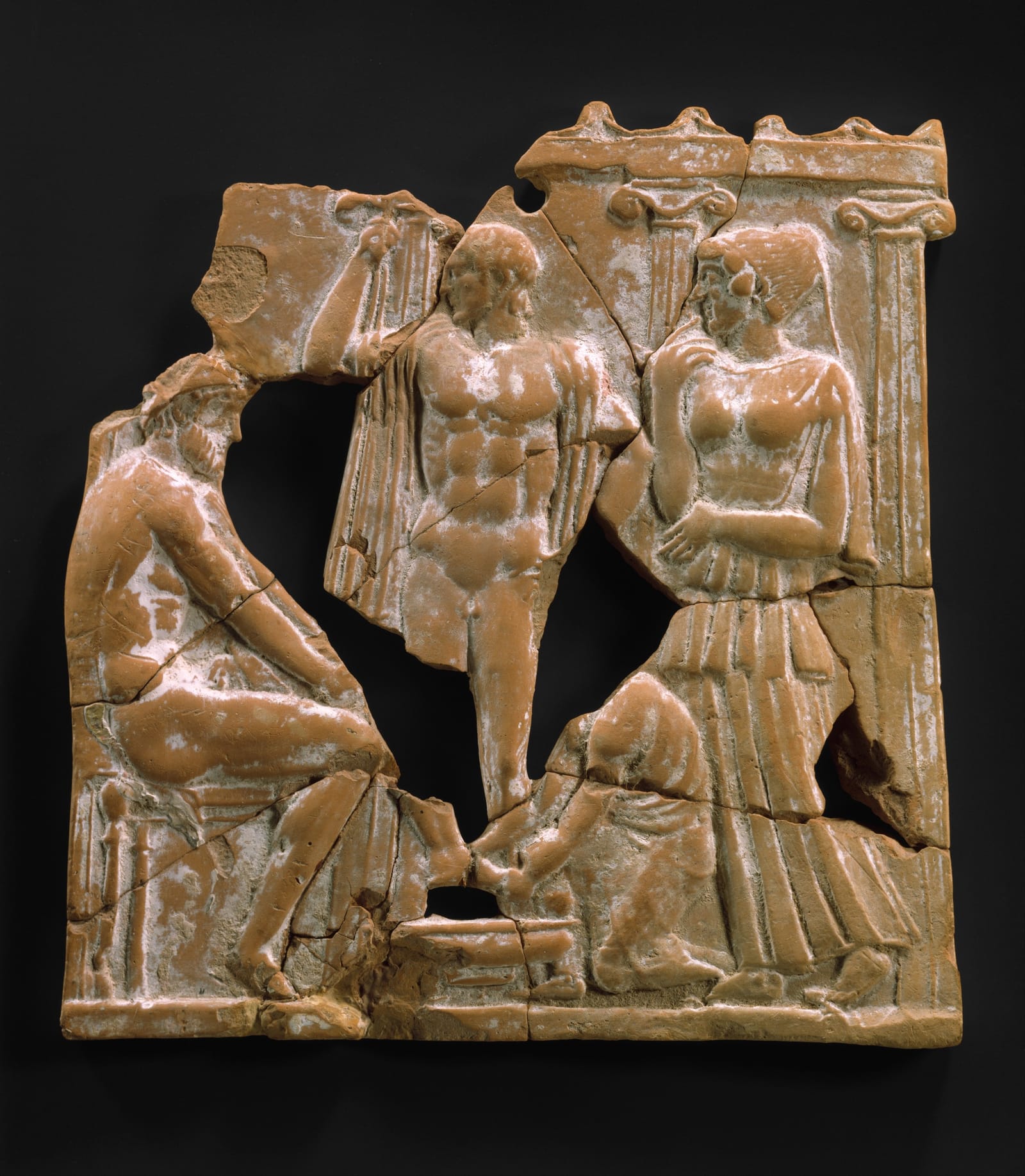 A broken clay relief plaque, pieced together: a bearded man sits on the left, a young man stands in the middle, and a woman in a long dress sits beside a column on the right.