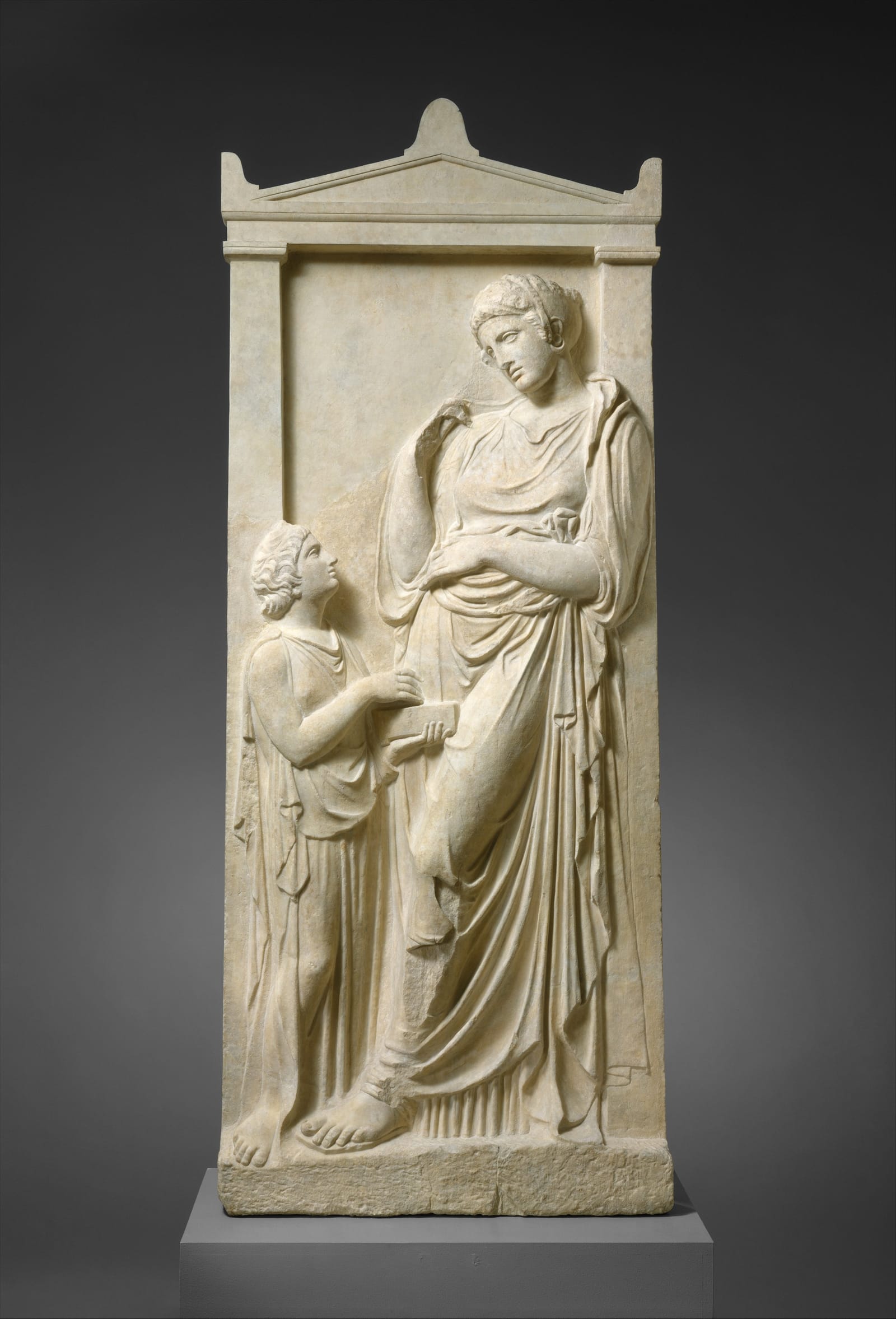 A tall marble gravestone topped with a small pediment: a young woman in a long draped dress stands looking down at a servant girl, carved at half her size, who holds up an open box to her.