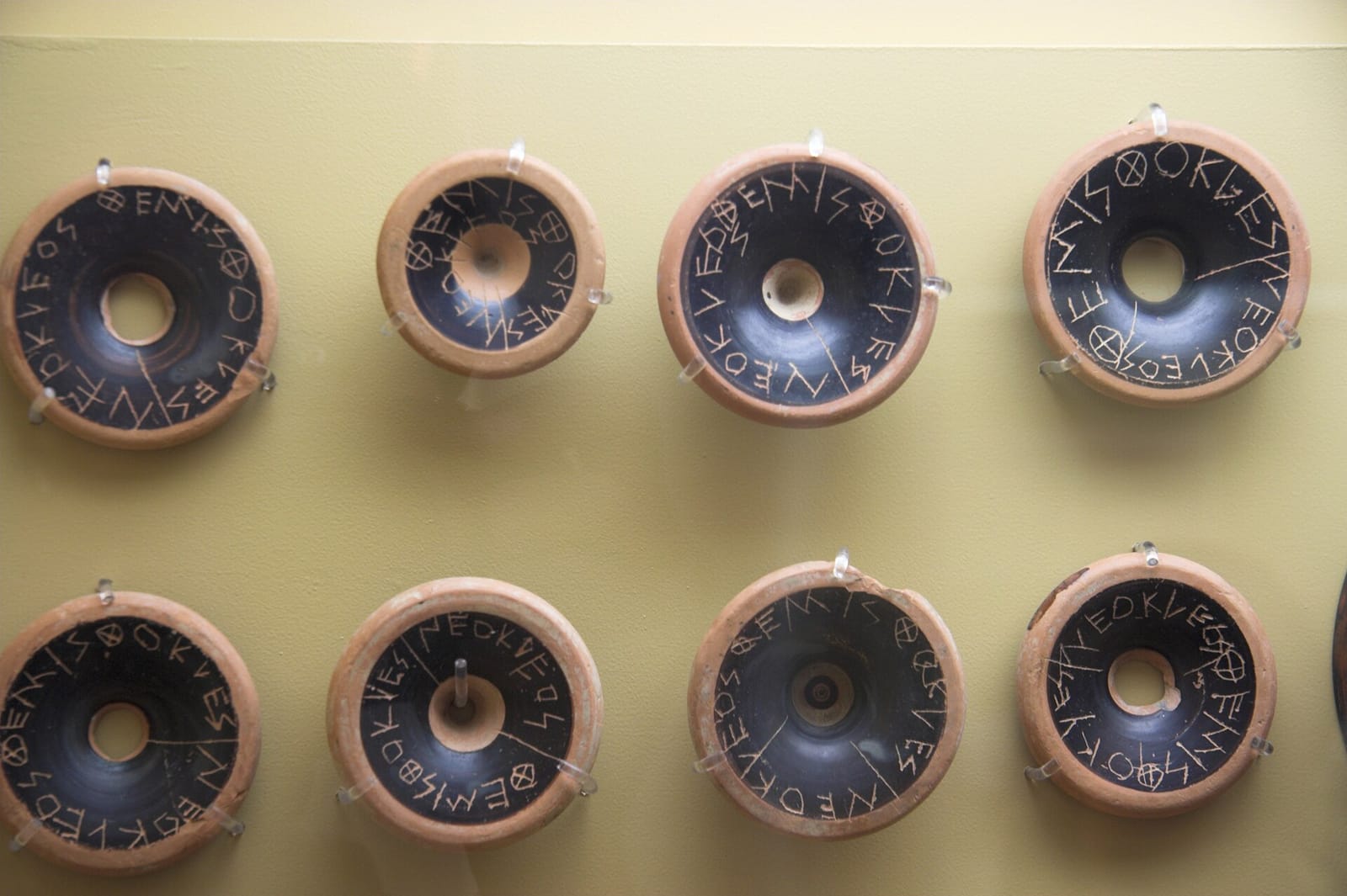 Round fragments of black-glazed pottery displayed on a wall, each scratched with a name in Greek letters.
