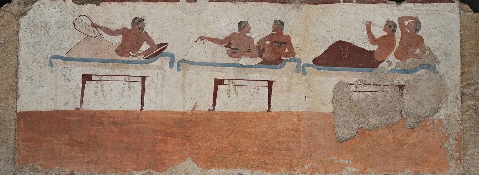 A painted tomb wall: young men reclining in pairs on couches, one of them flicking the last drops from his cup.